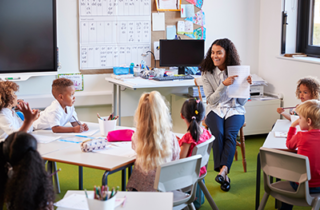 Image of a female teacher sitting in front of a class with 7 children looking at a paper in her hands that she's pointing to.