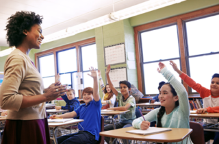 A teacher faces a group of students who are raising their hands.