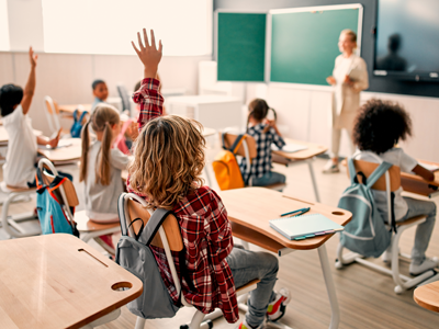 A class of kids raise their hands in front of their teacher.